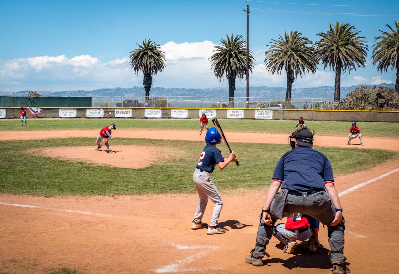 Children playing baseball in an outdoor field under a bright blue sky with palm trees in the background.