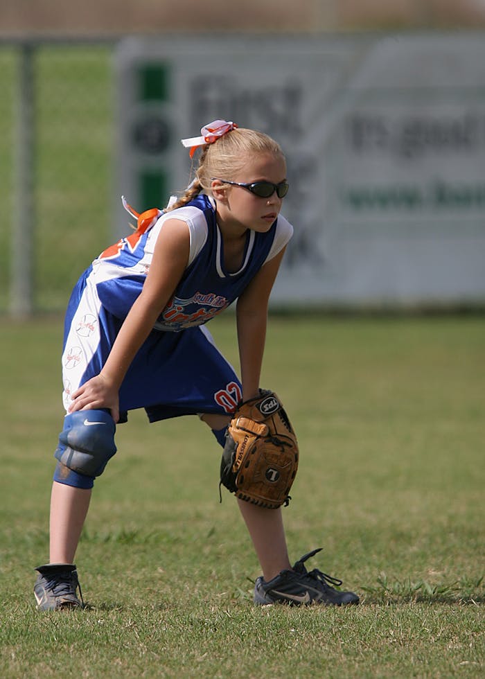 A young girl in a softball uniform and sunglasses focuses during a game.